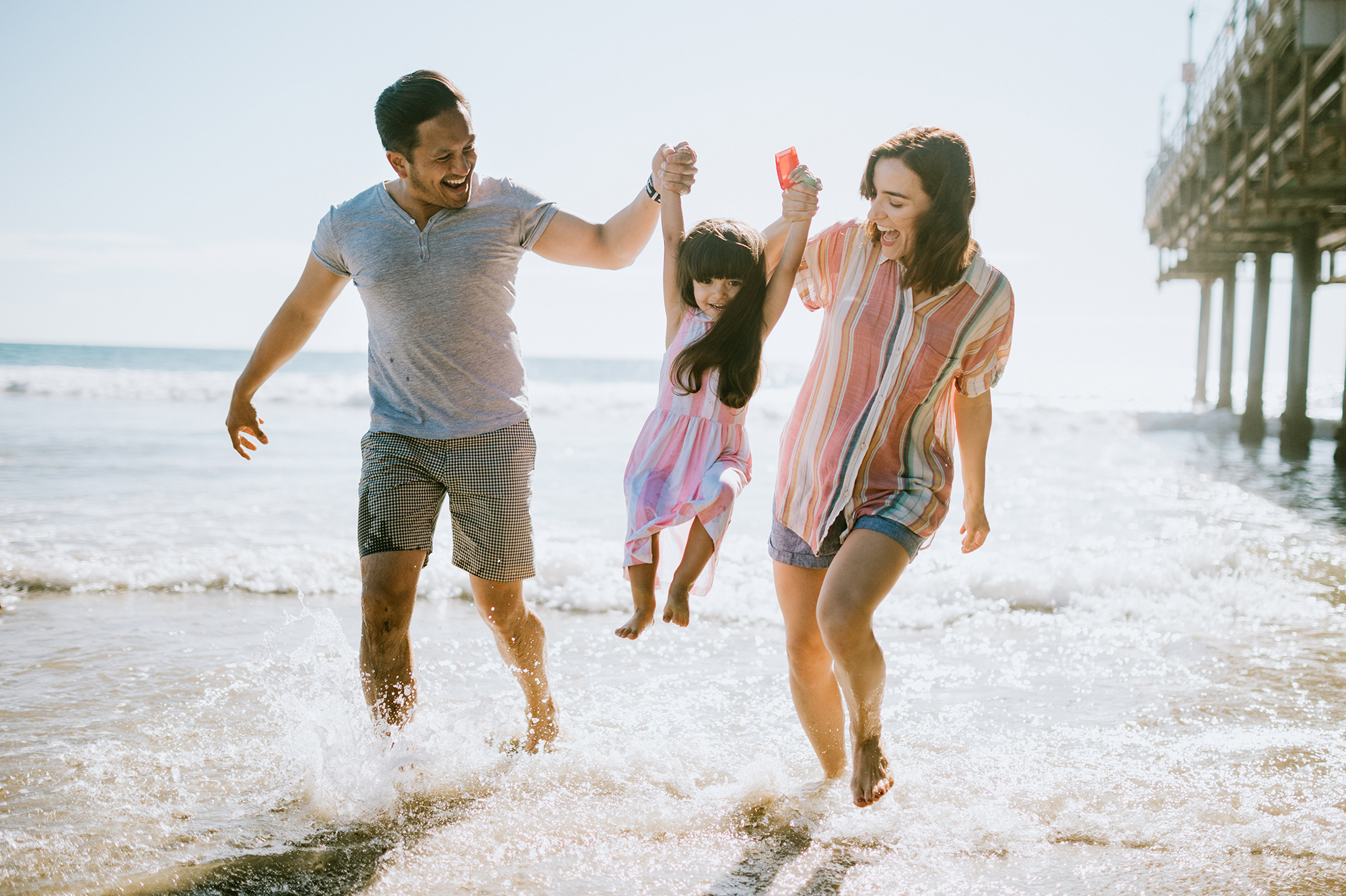 Family of three walking in surf next to a pier.