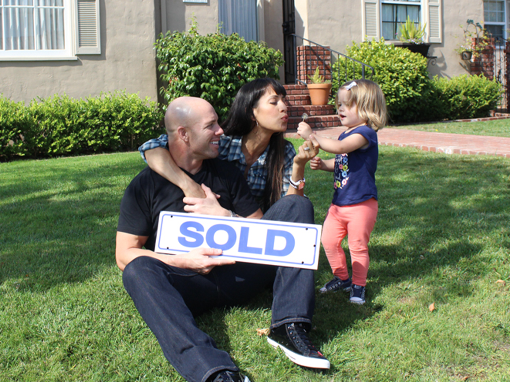 A family sits in their front yard, holding a "sold" sign